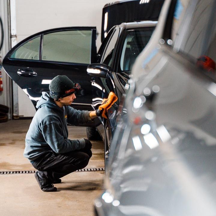 A person kneels, wiping a black car with an orange cloth inside a car detailing shop. Another car is visible in the foreground.