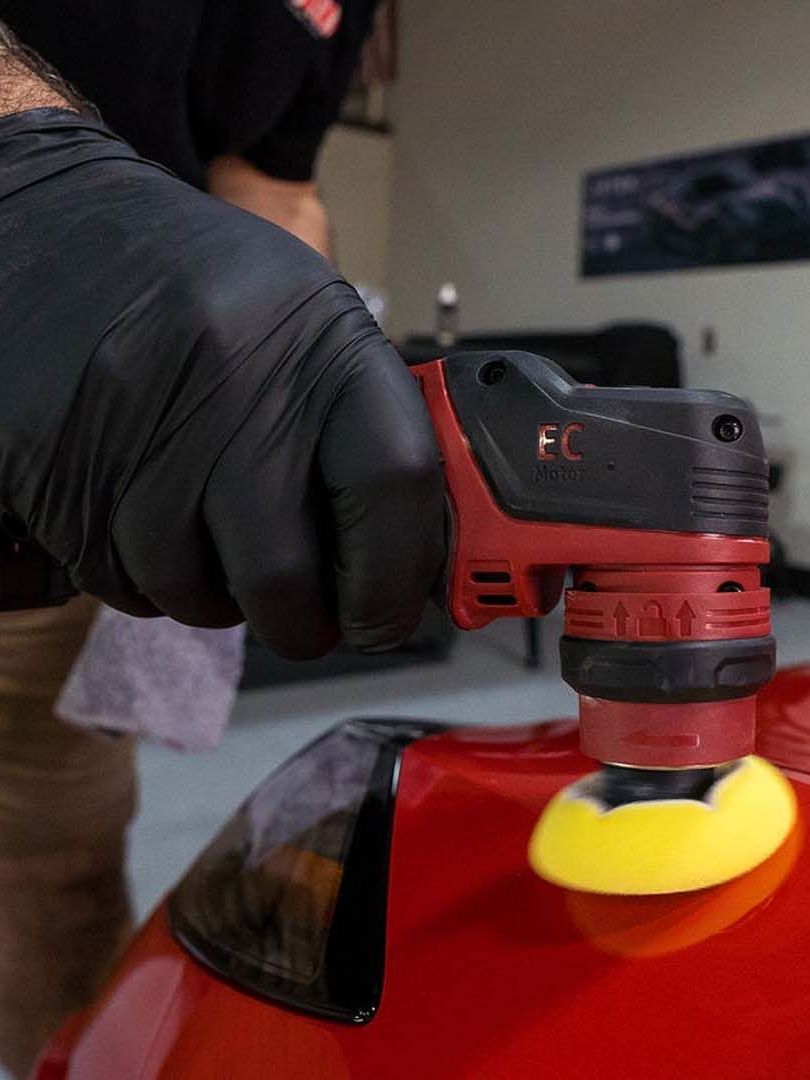 A gloved hand polishes a red car with a yellow buffing pad and power tool. The background is blurred.