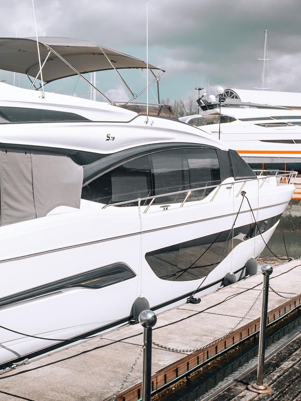 White motor yacht docked at a marina on a cloudy day. Another yacht is partially visible in the background.