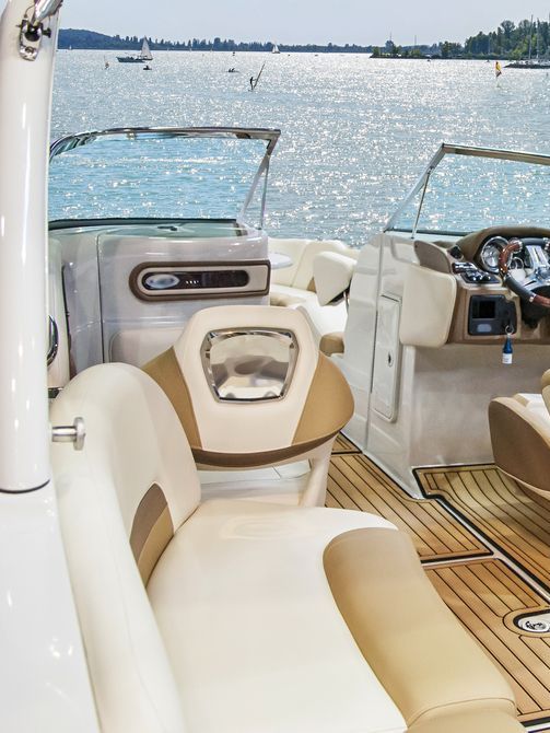 Interior of a boat with beige and brown seating, wooden flooring, and a view of the water and shoreline.