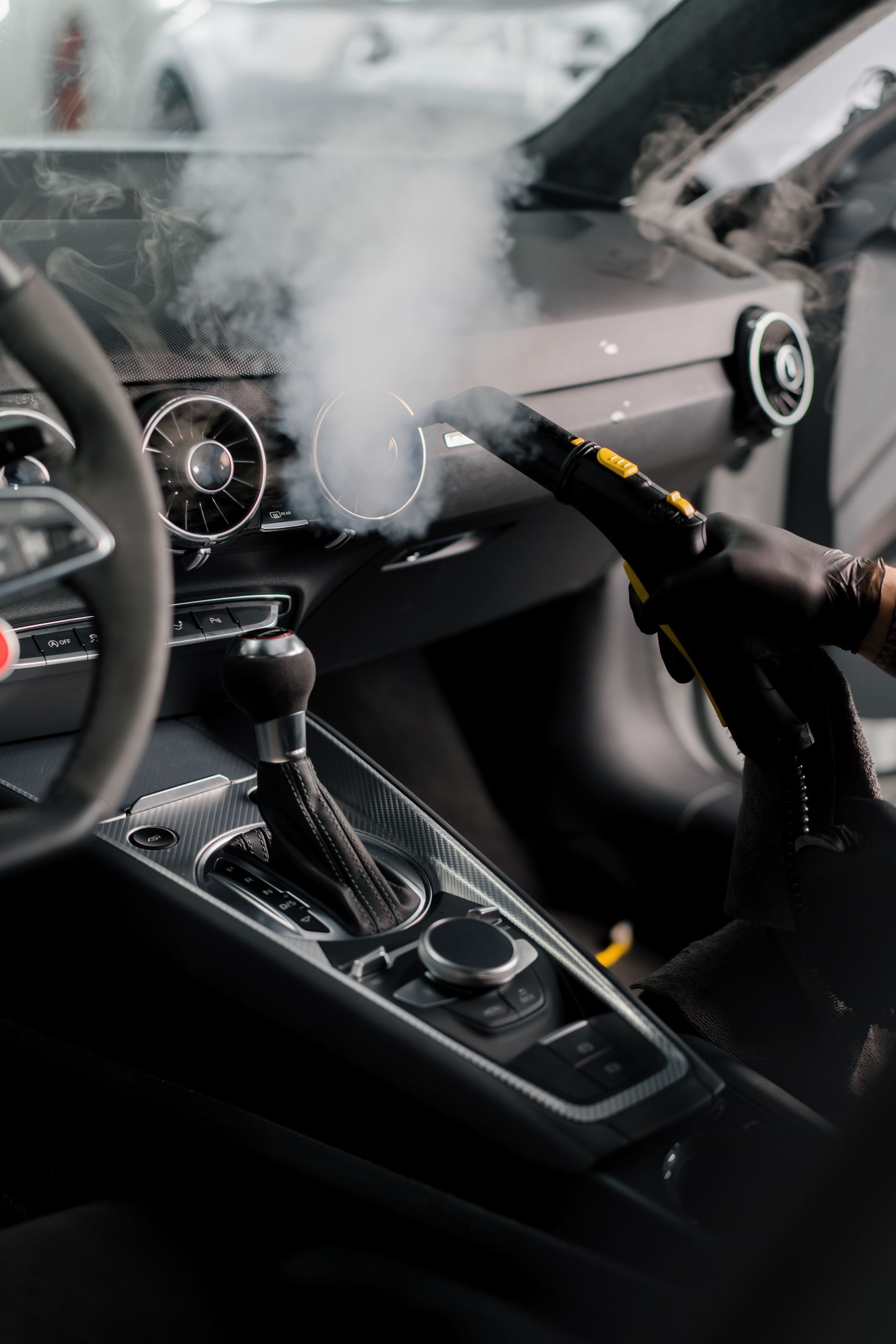 A gloved hand using a steam cleaner on a car's dashboard, removing dirt.