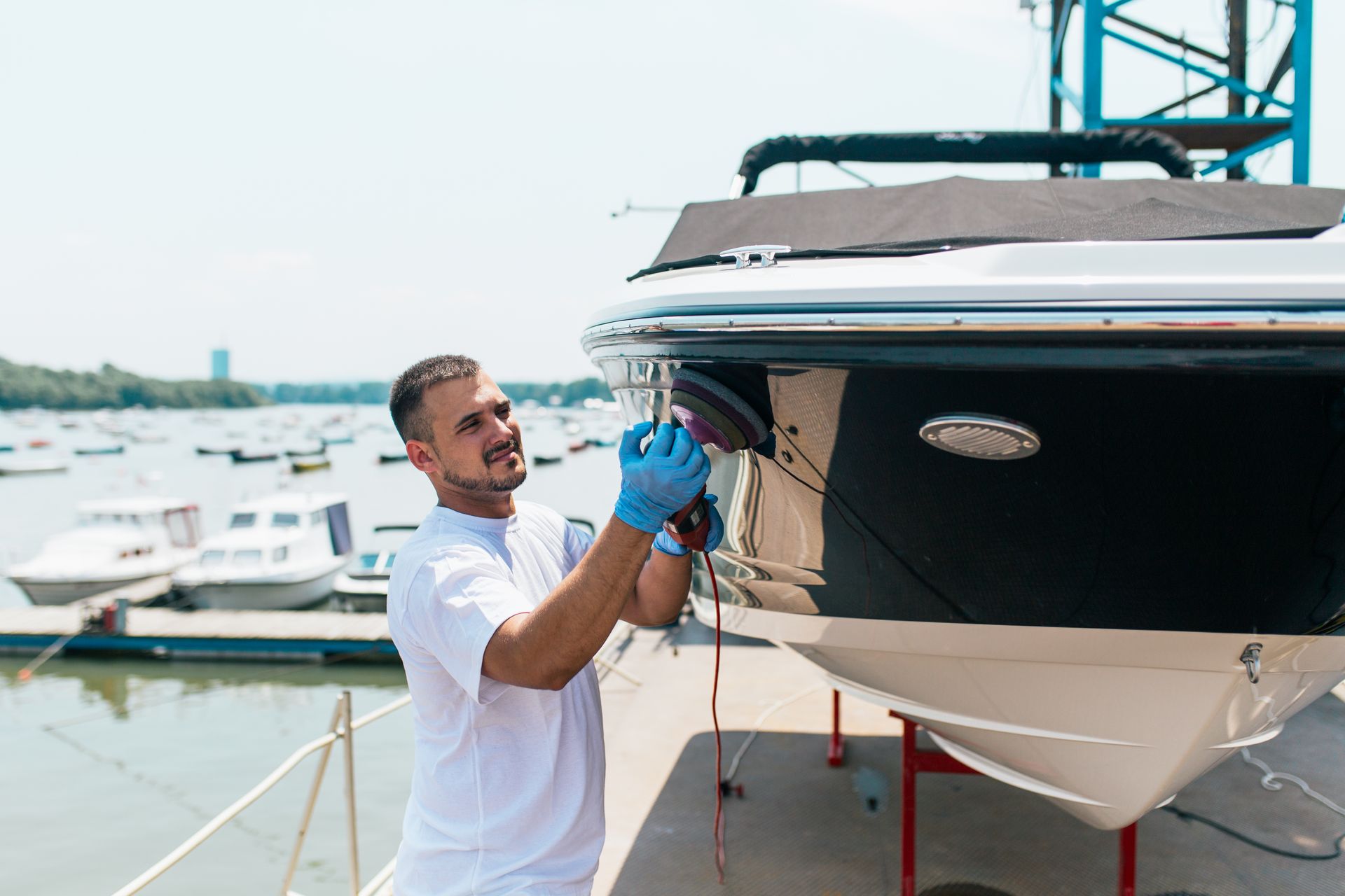 A man polishes a black and white boat at a marina, wearing gloves and a white shirt.