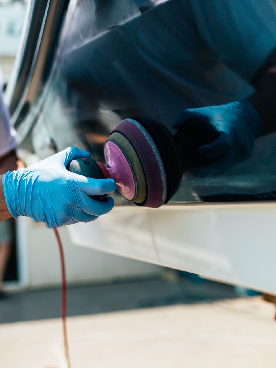 Person in blue gloves polishing a white boat with a power buffer. Outdoors, with bright sunlight.