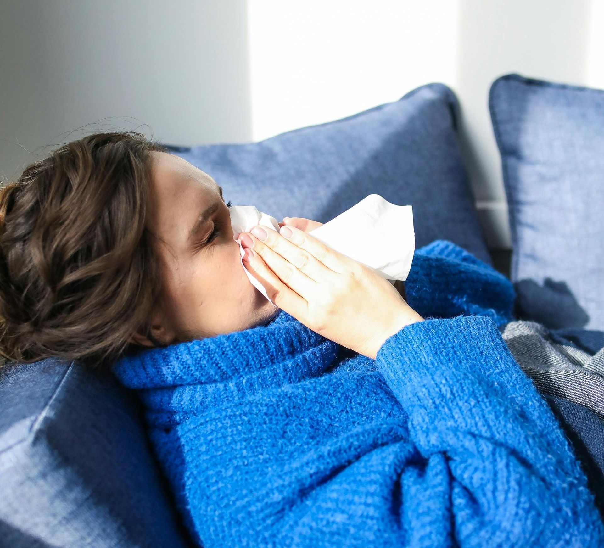A woman is laying on a couch blowing her nose with a napkin.