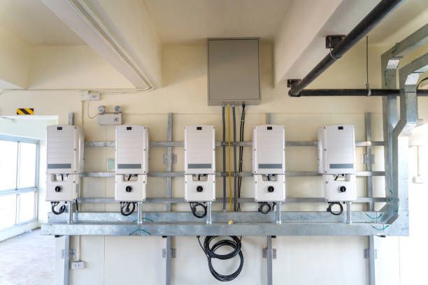 Five white solar inverters mounted on a wall, connected to wiring in an indoor setting.