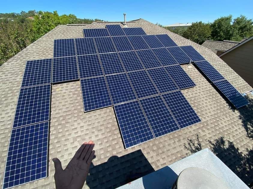Solar panels on a shingled roof under a blue sky; person's hand in shadow.