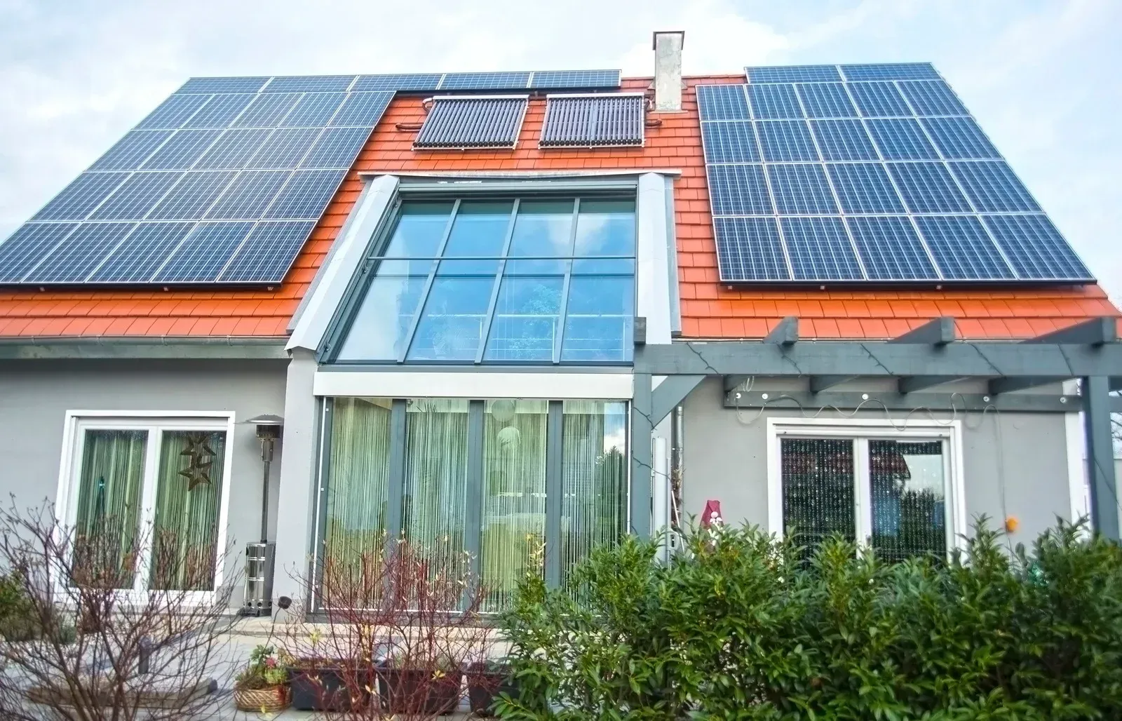 House with red roof, solar panels, and large glass window. Green bushes and gray exterior walls.