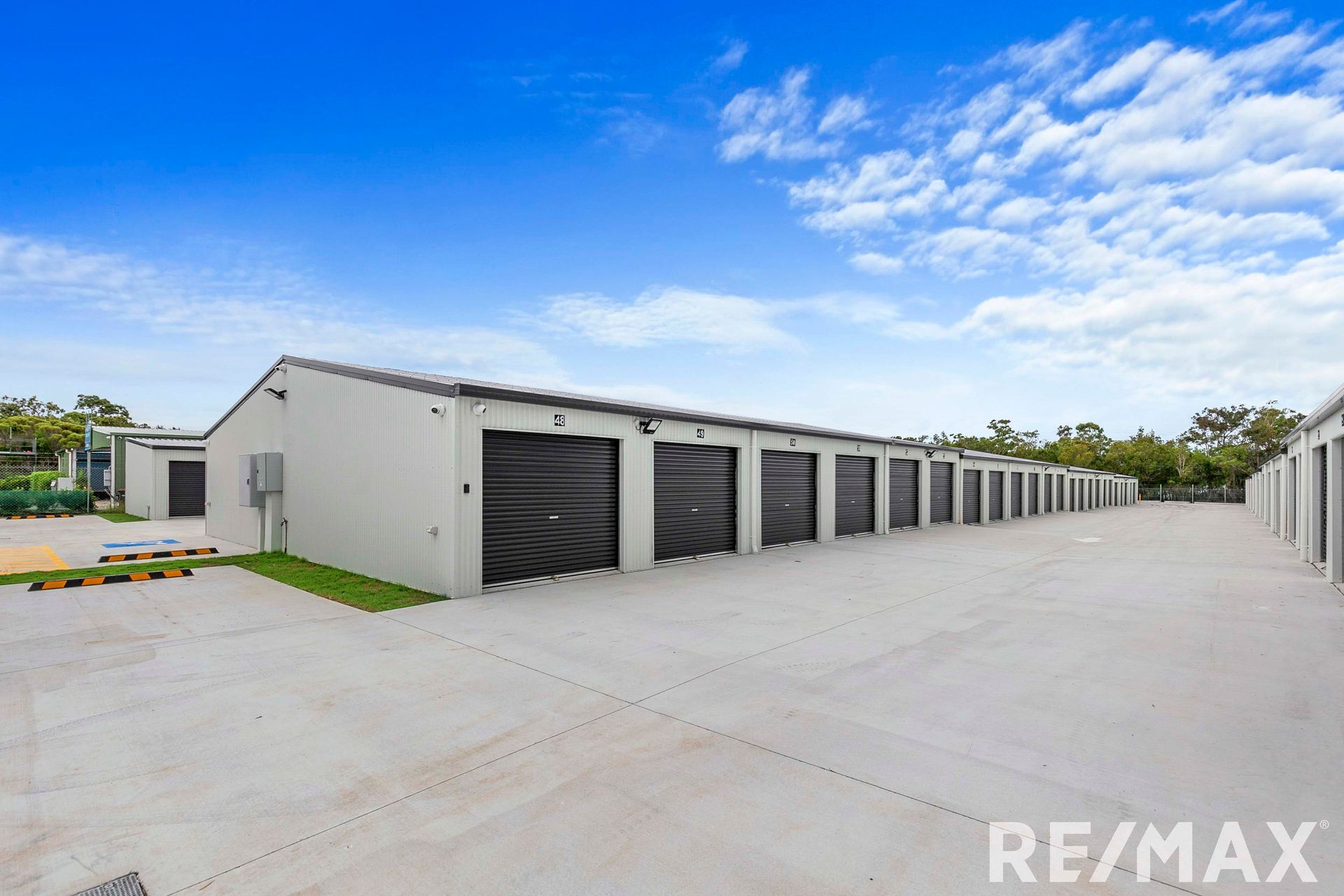 Exterior view of a storage facility with multiple units. Gray roll-up doors, concrete lot, and blue sky.