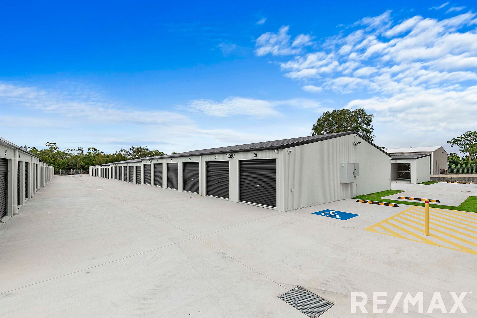 Exterior view of a storage facility with multiple units. Gray buildings, black doors, concrete ground, blue sky.