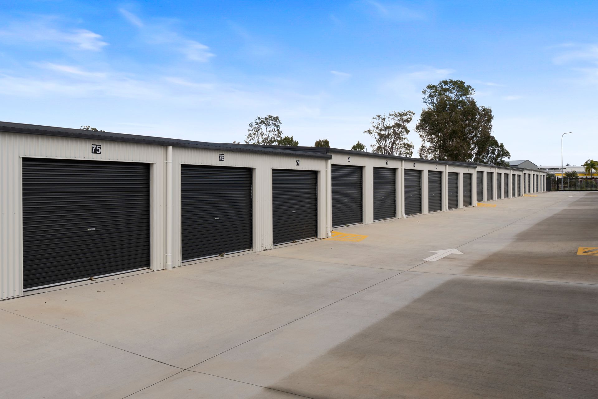 Exterior view of a storage facility with multiple units. Gray roll-up doors, concrete lot, and blue sky.