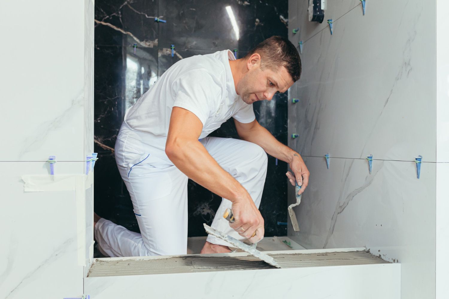 A man is kneeling down while laying tiles in a bathroom.