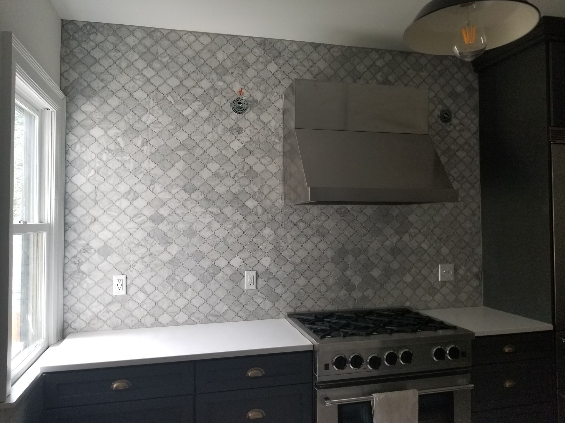 Kitchen with gray cabinets, white countertops, and fish-scale tile backsplash. Stainless steel range hood and stove.