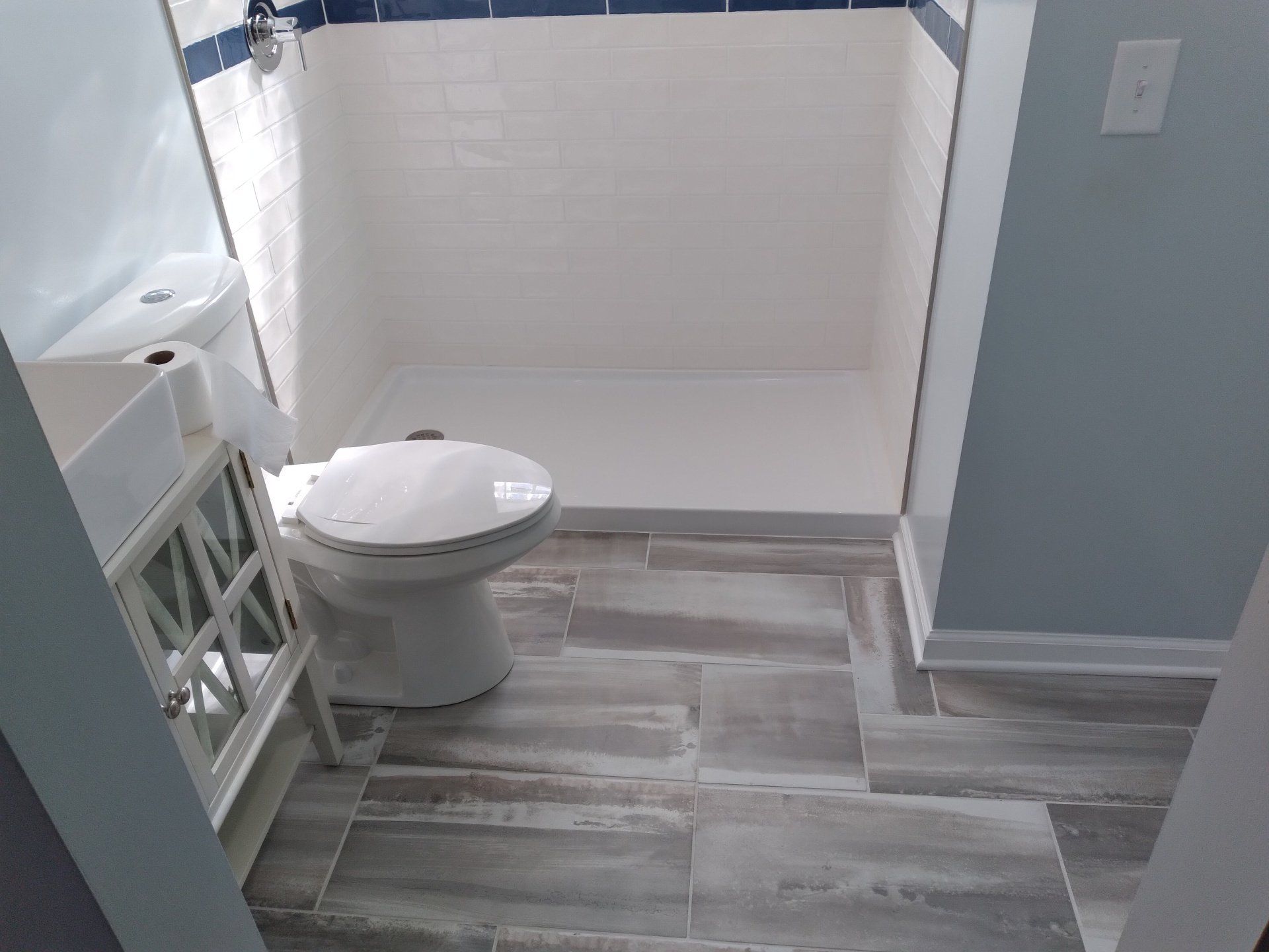 Bathroom with white shower and toilet, wood-look floor tiles, white vanity, and light blue walls.