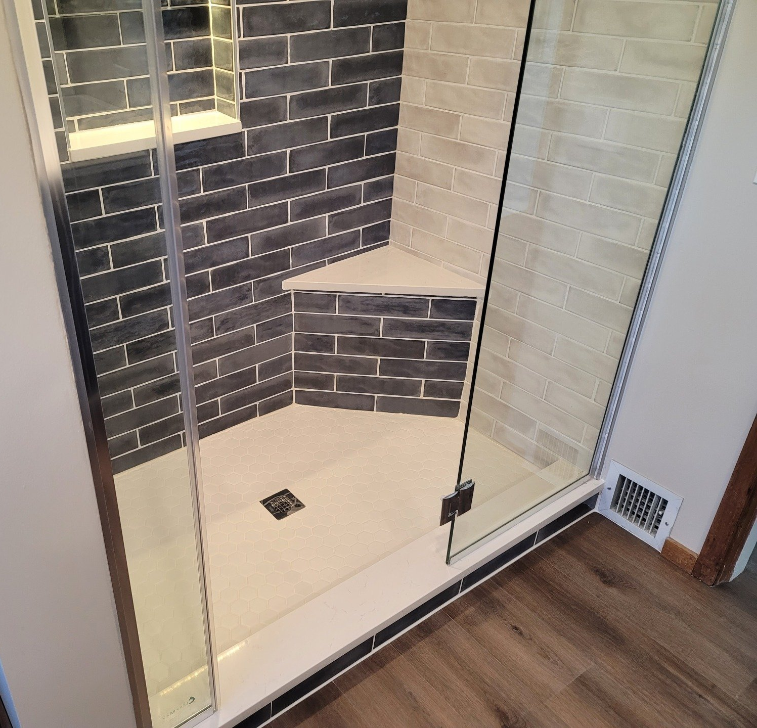 Shower with glass door, black and beige tile, built-in seat, and light wood flooring.