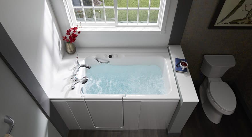White walk-in bathtub with open door, filled with water, next to toilet, under window with flowers.