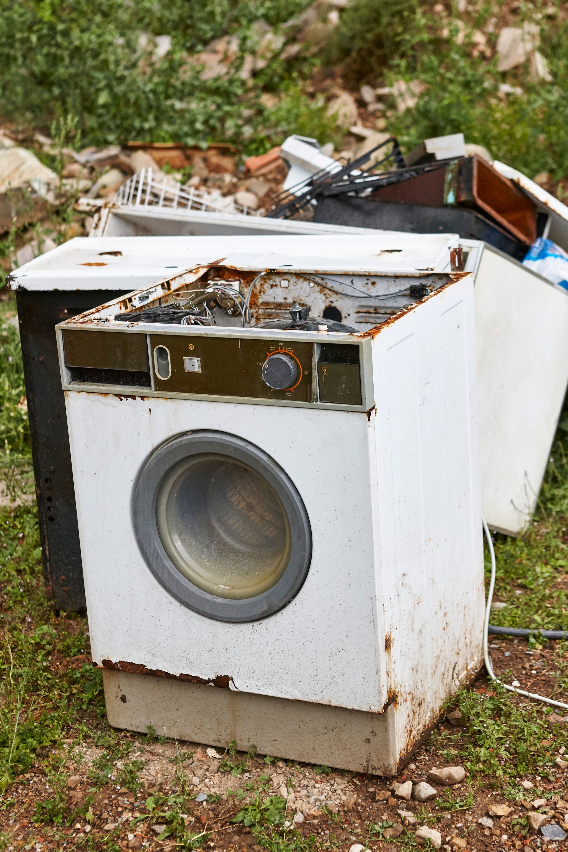 An old washing machine is sitting in the grass next to a pile of junk.