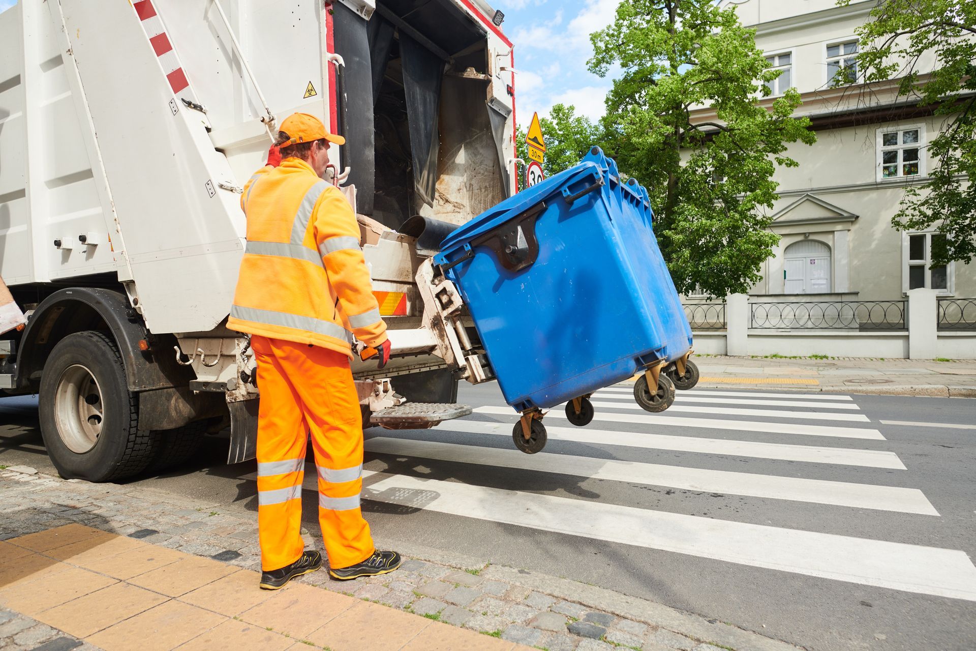 A garbage man is loading a blue trash can into a garbage truck.
