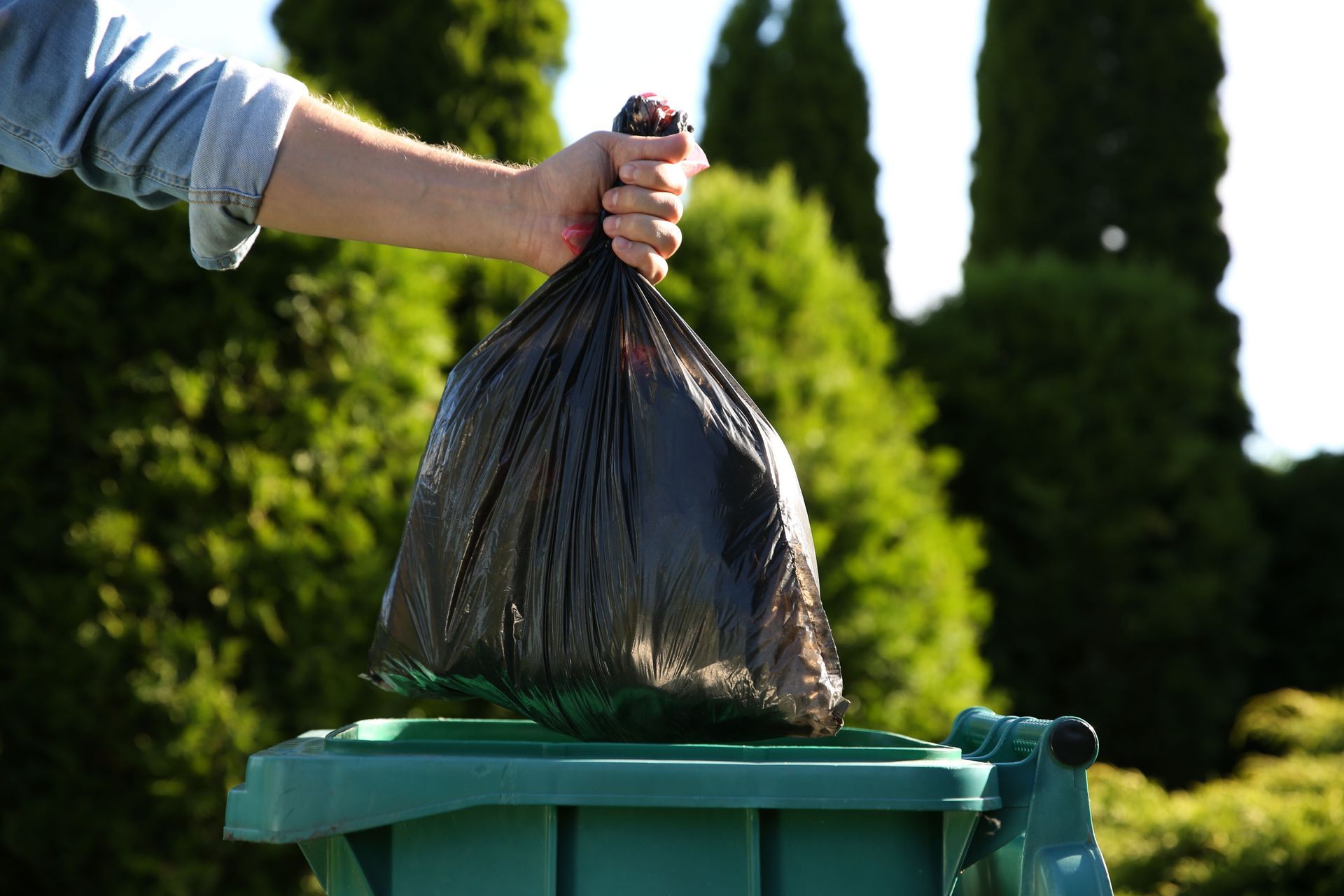 A person is putting a black trash bag into a green trash can.