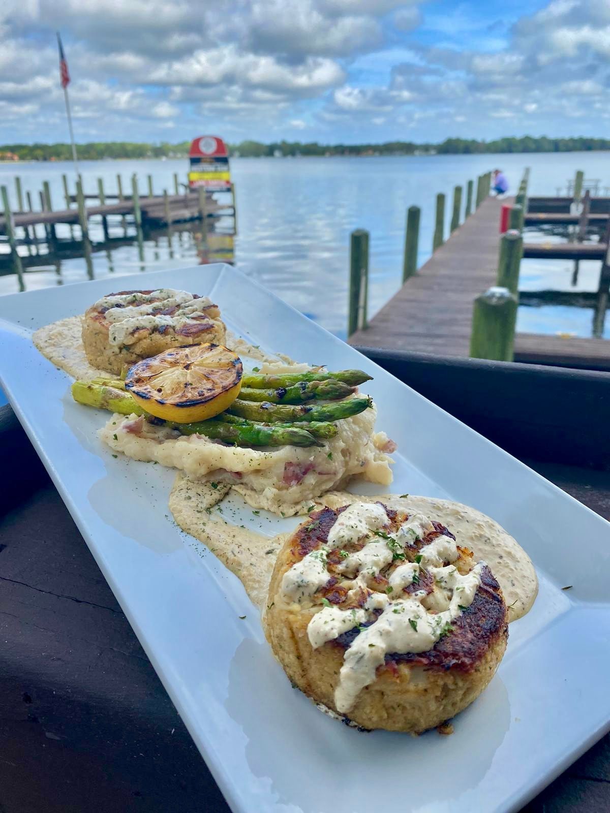 A rectangular white plate with two crab cakes, asparagus, and a lemon slice, set against a lakeside dock view.