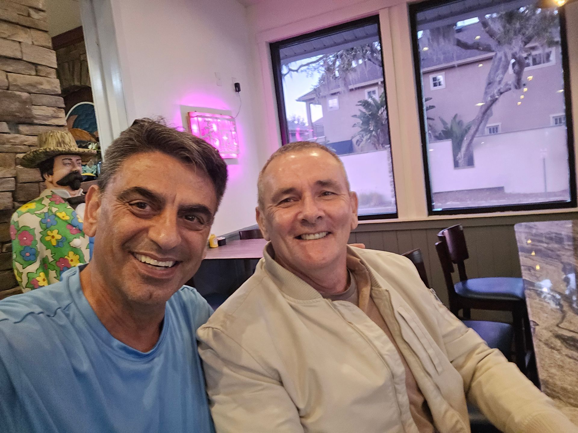 Two men smiling while taking a selfie together in a brightly lit indoor restaurant with windows in the background.