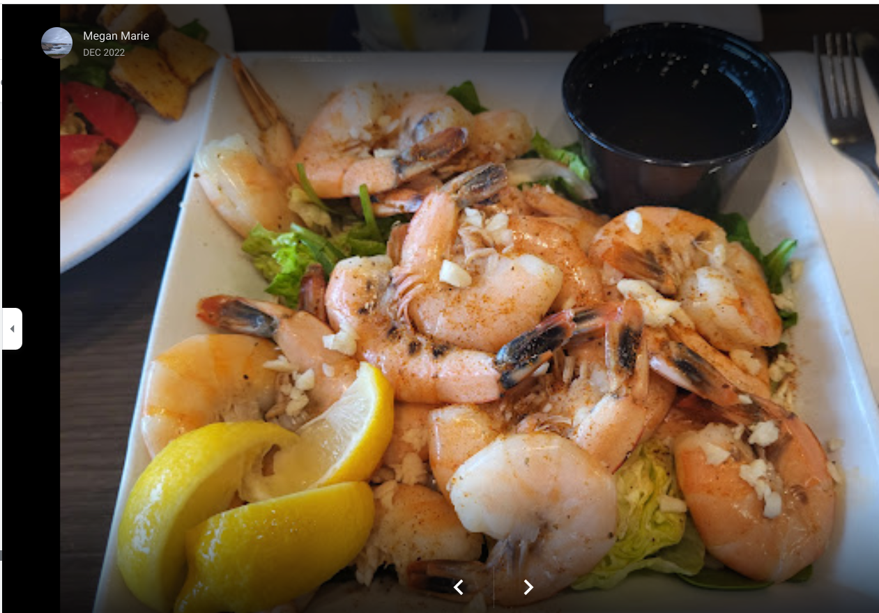A rectangular plate of cooked, garlicky shrimp served over greens with two lemon wedges and a side of dipping sauce.