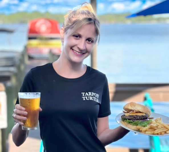 A server in a black Tarpon Turtle t-shirt smiles while holding a glass of beer and a burger with fries by the water.