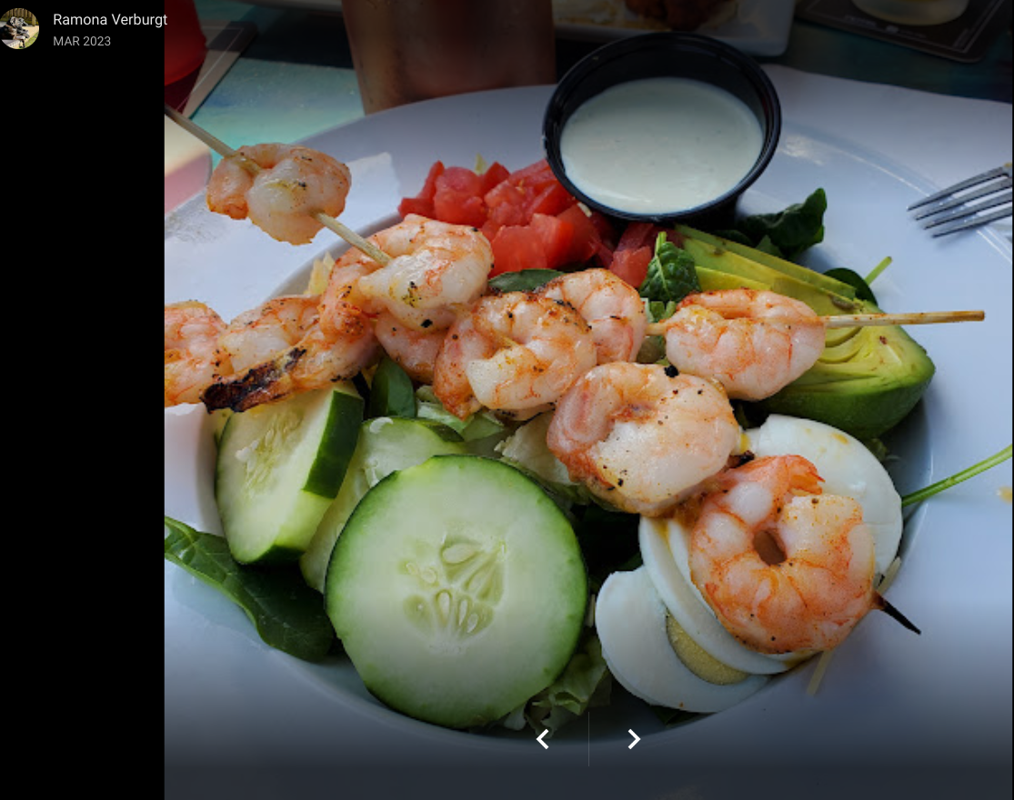 A bowl of salad topped with shrimp skewers, sliced cucumbers, tomatoes, avocado, a hard-boiled egg, and a side of dressing.