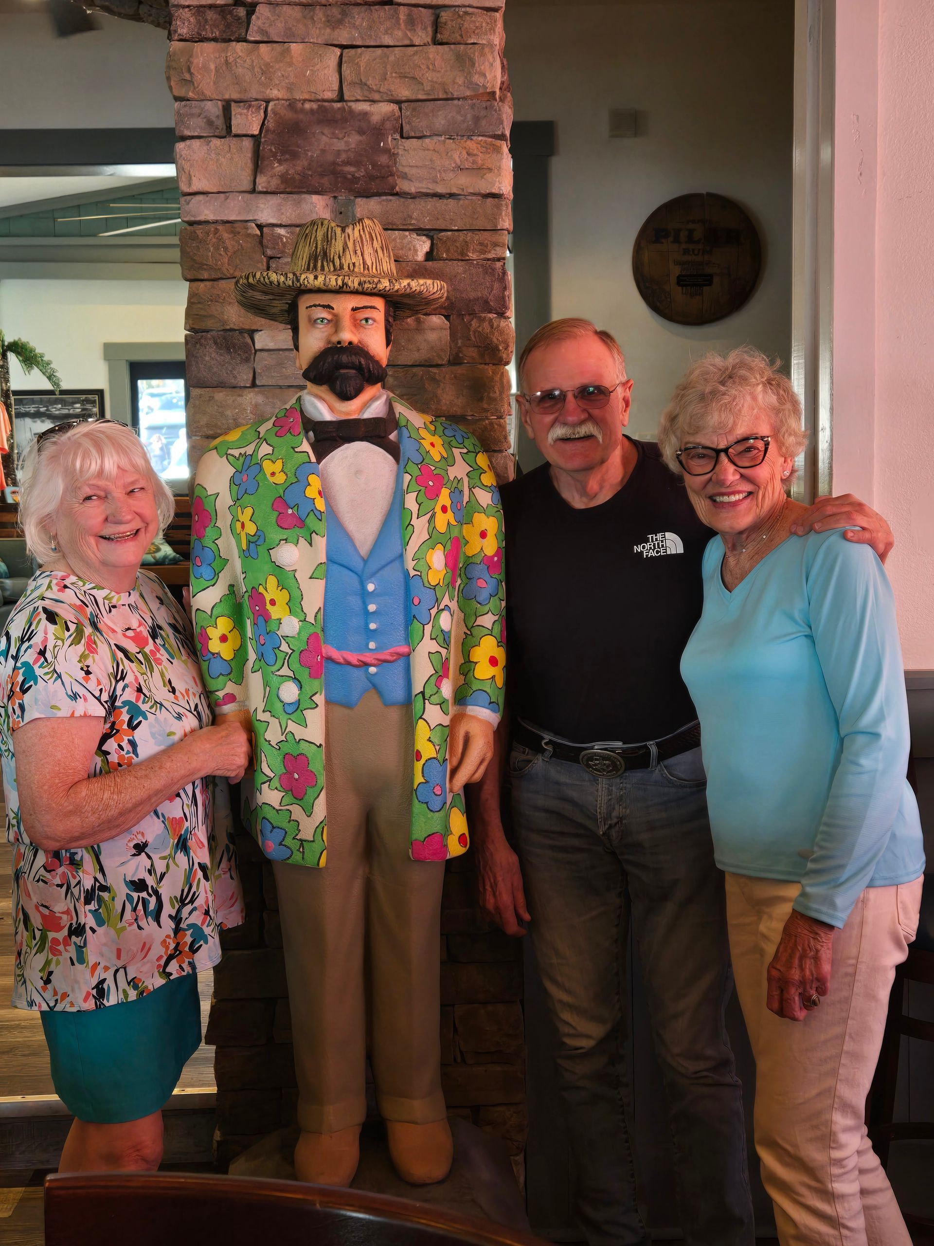 Three people stand smiling for a photo next to a colorful, patterned statue of a man in a hat near a stone pillar.