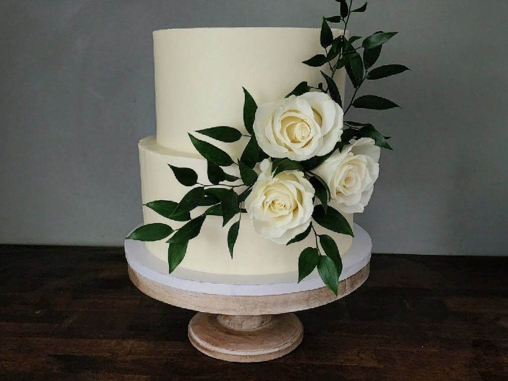 A white wedding cake with white roses and green leaves on a wooden table.