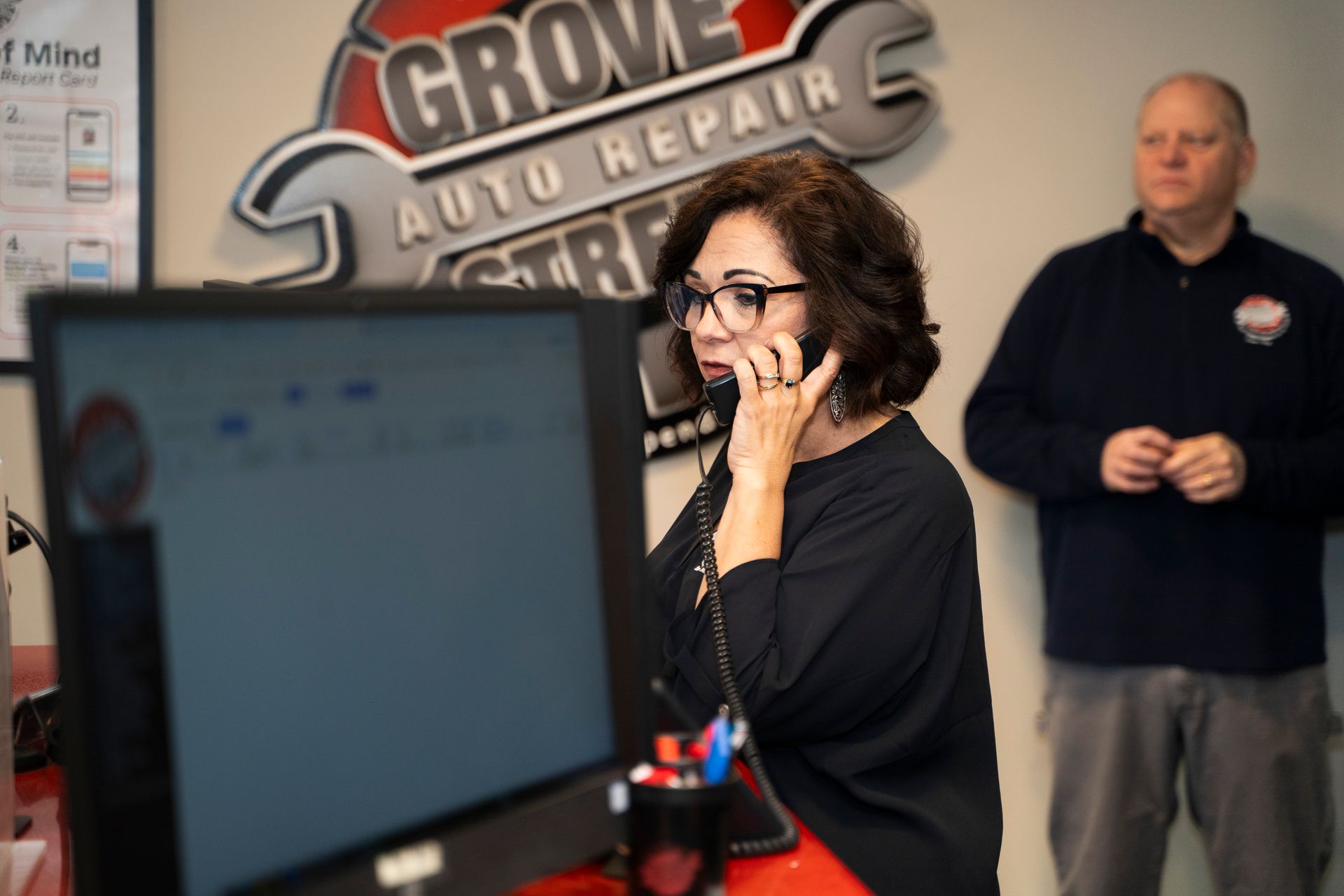 Woman on phone at computer, inside Grove Auto Repair; man stands behind her.
