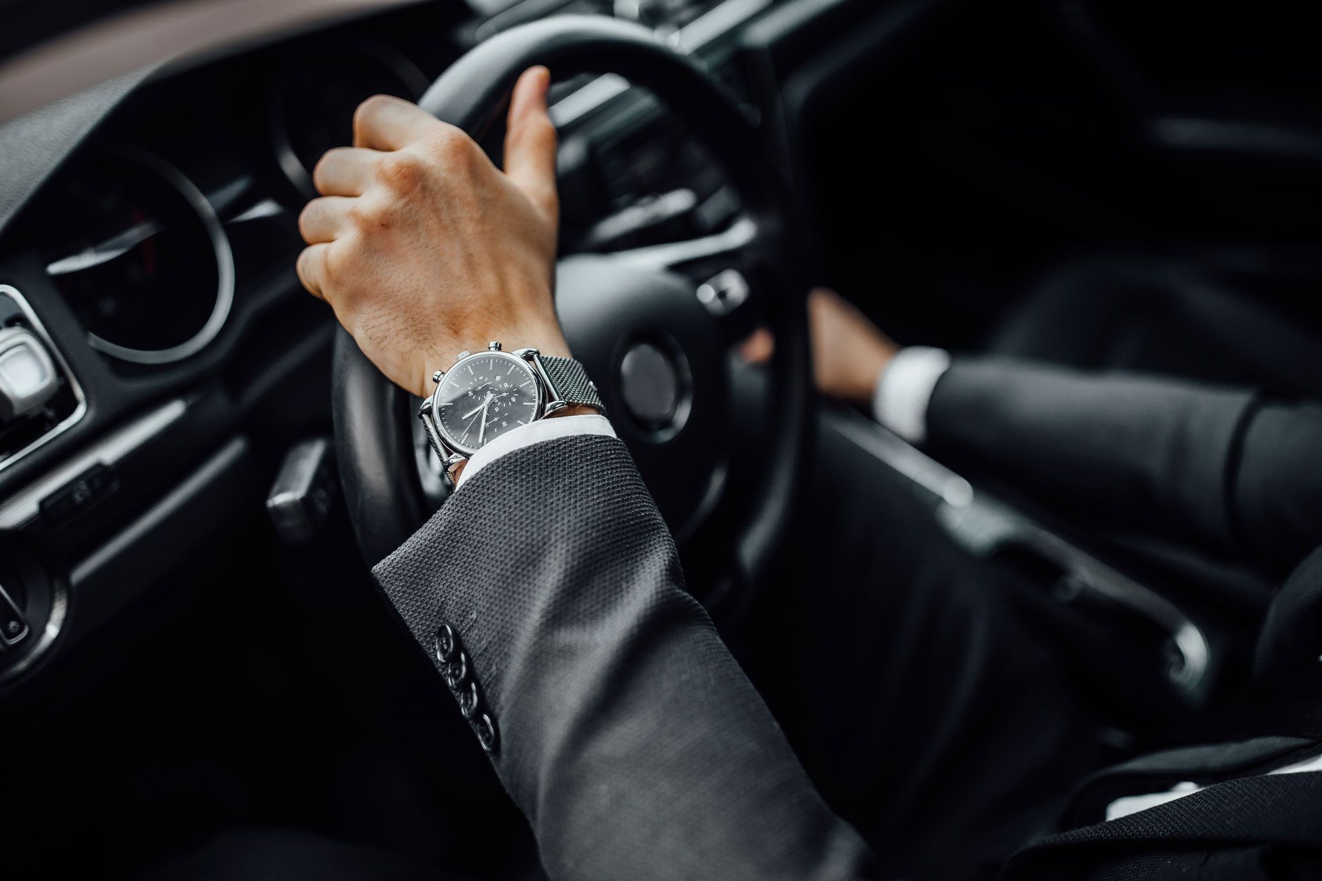 Driver’s hand on steering wheel inside a car, wearing a silver watch and dark suit sleeve