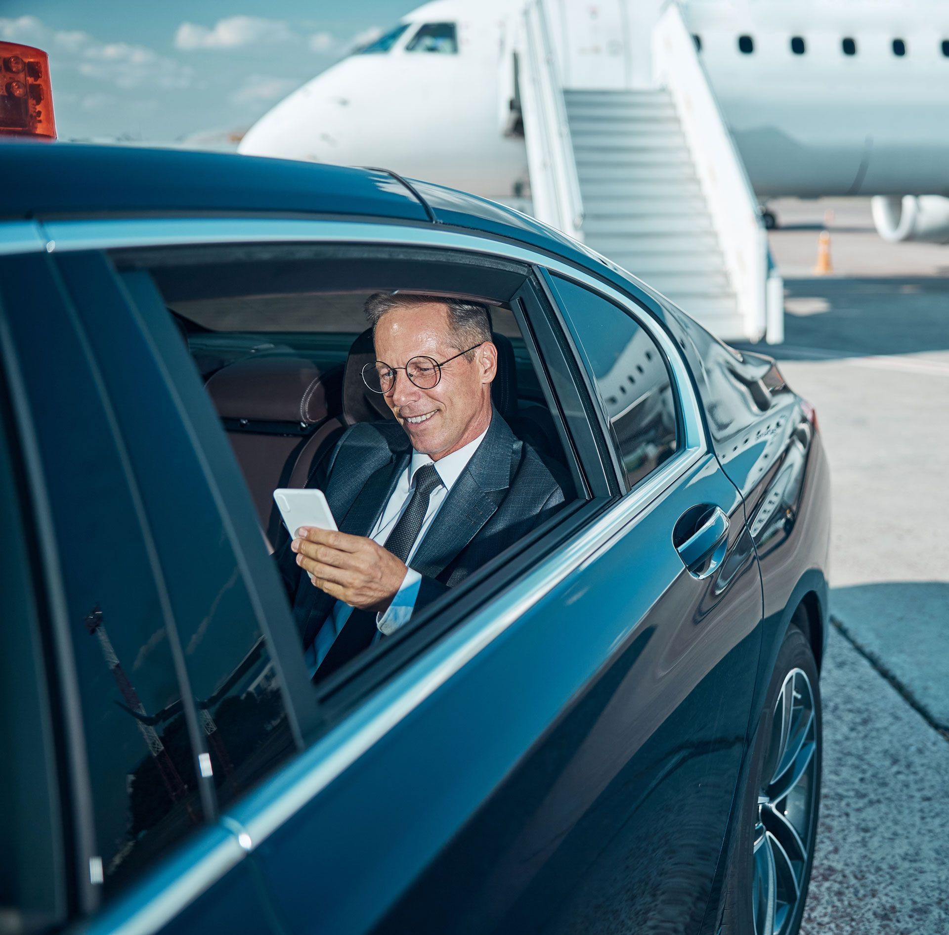 Man in a blue car smiling at a phone near an airport boarding stairway