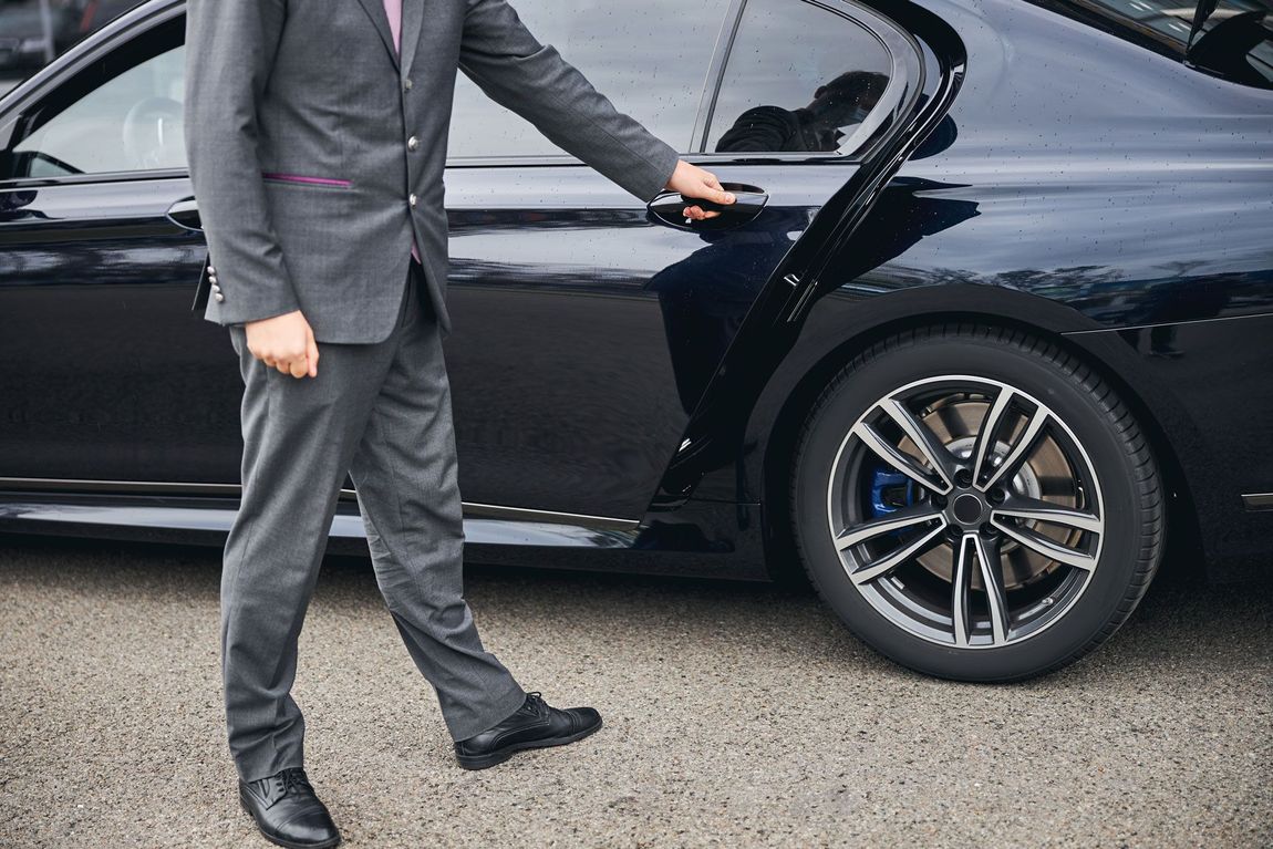 Person in a gray suit opening the door of a black luxury car.