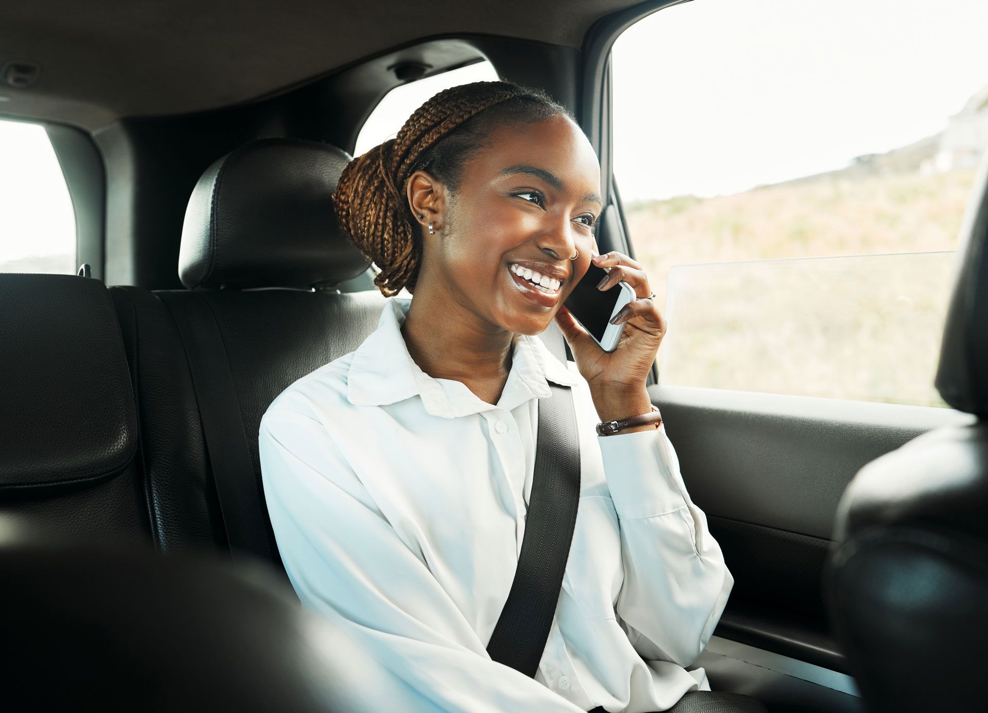 Woman smiling and talking on a phone in the back seat of a car, wearing a white shirt and seat belt