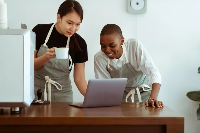 Two women in aprons look at laptop, one holding a mug. Indoors at a cafe.