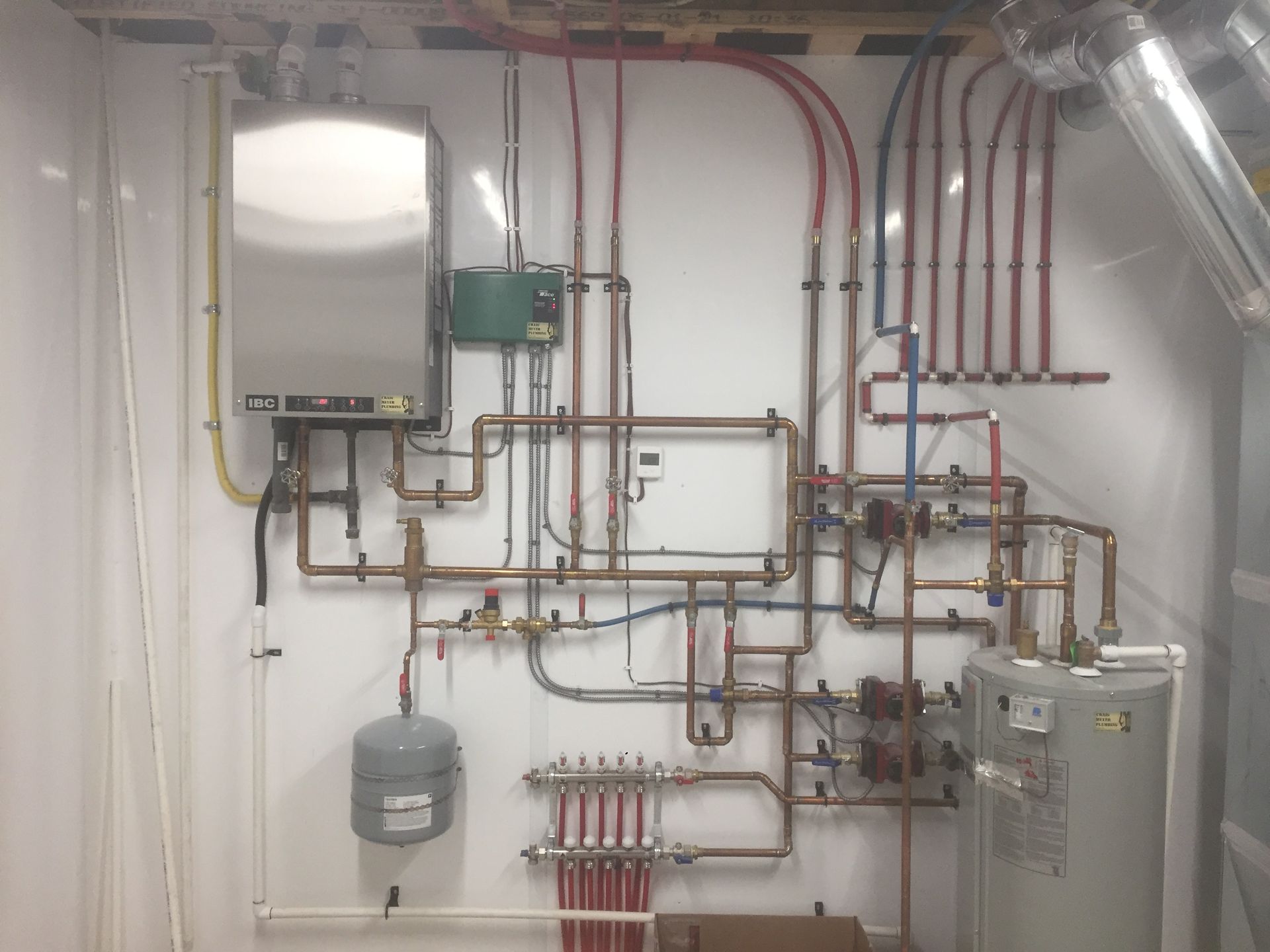Heating system with copper and colored piping, water heater, tank, and boiler, mounted on a white wall.