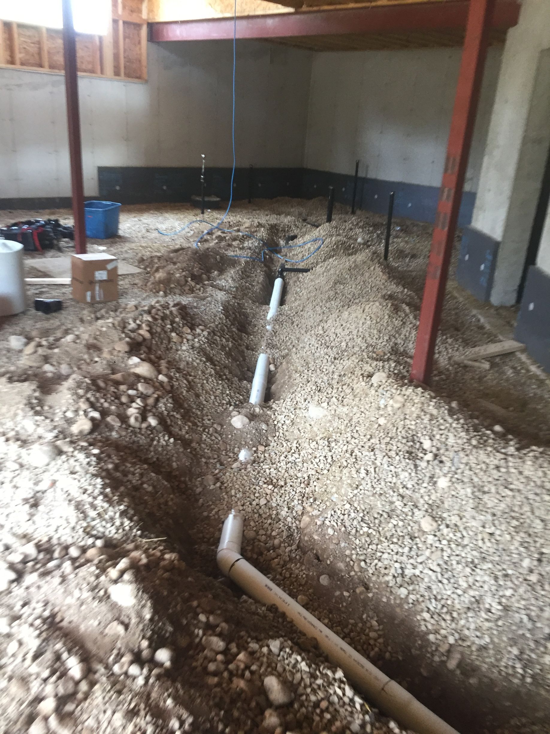 Basement with plumbing pipes in a trench. Gravel, dirt, and support beams are also visible.