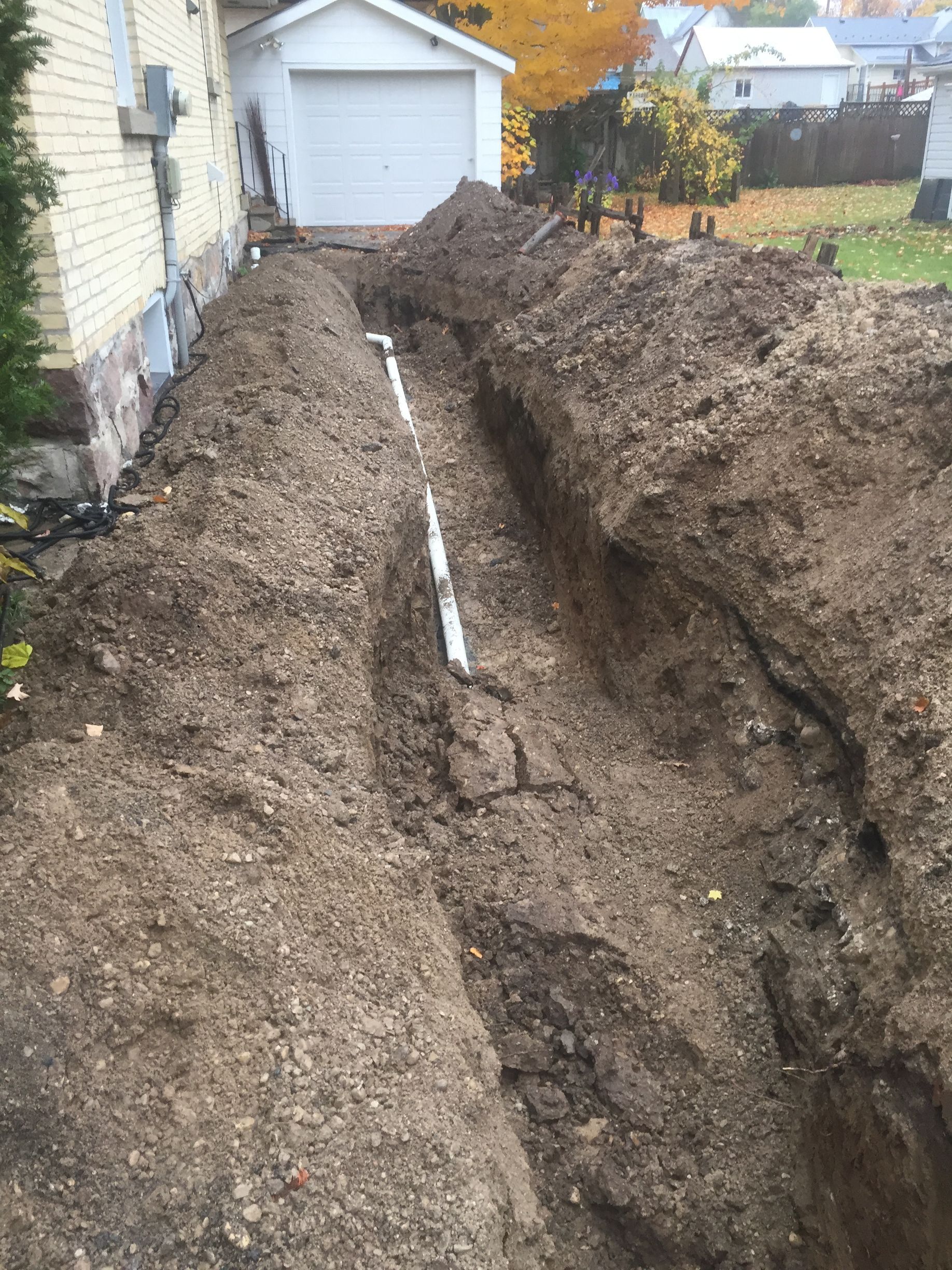 A long trench with a white pipe installed along a house and garage. Dirt surrounds.