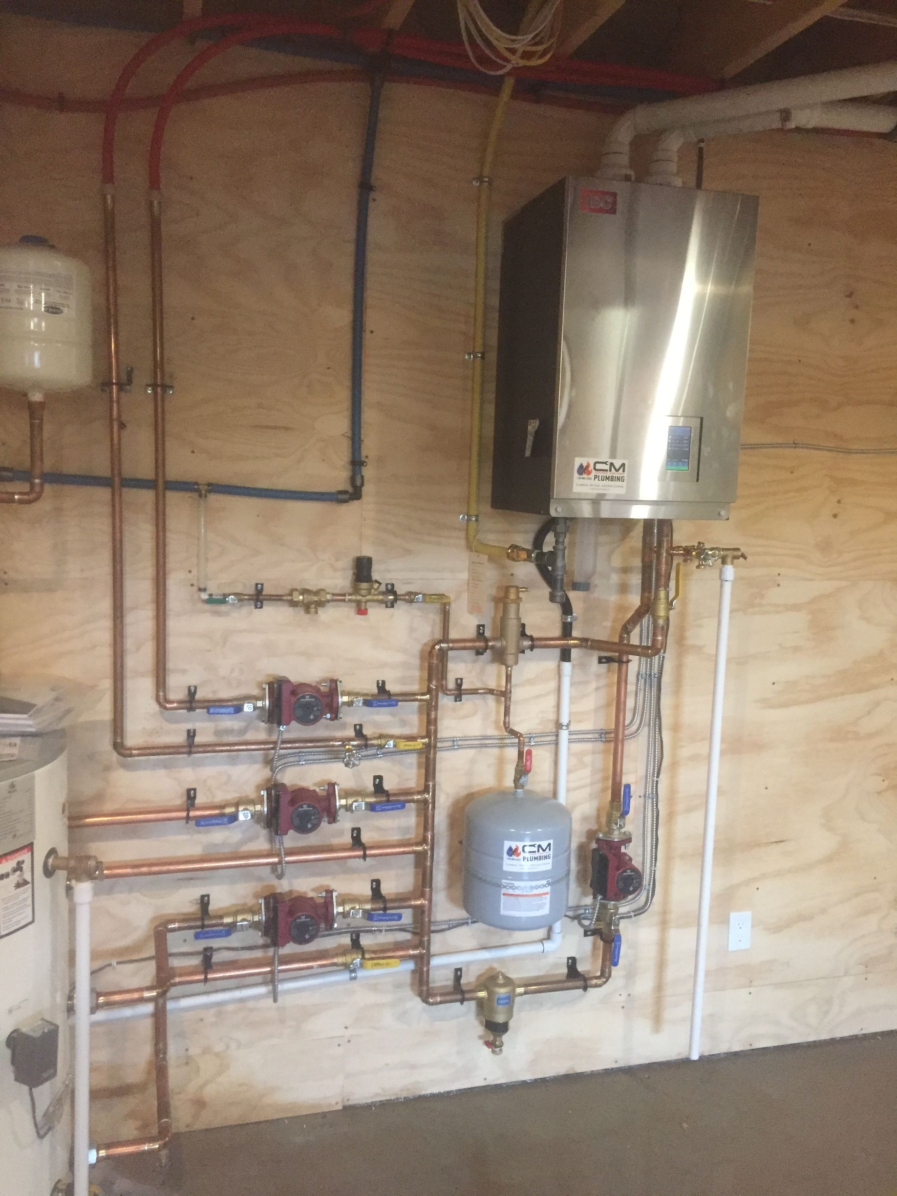 Copper pipes and tankless water heater installed on plywood wall. Various valves and a pressure tank are visible.