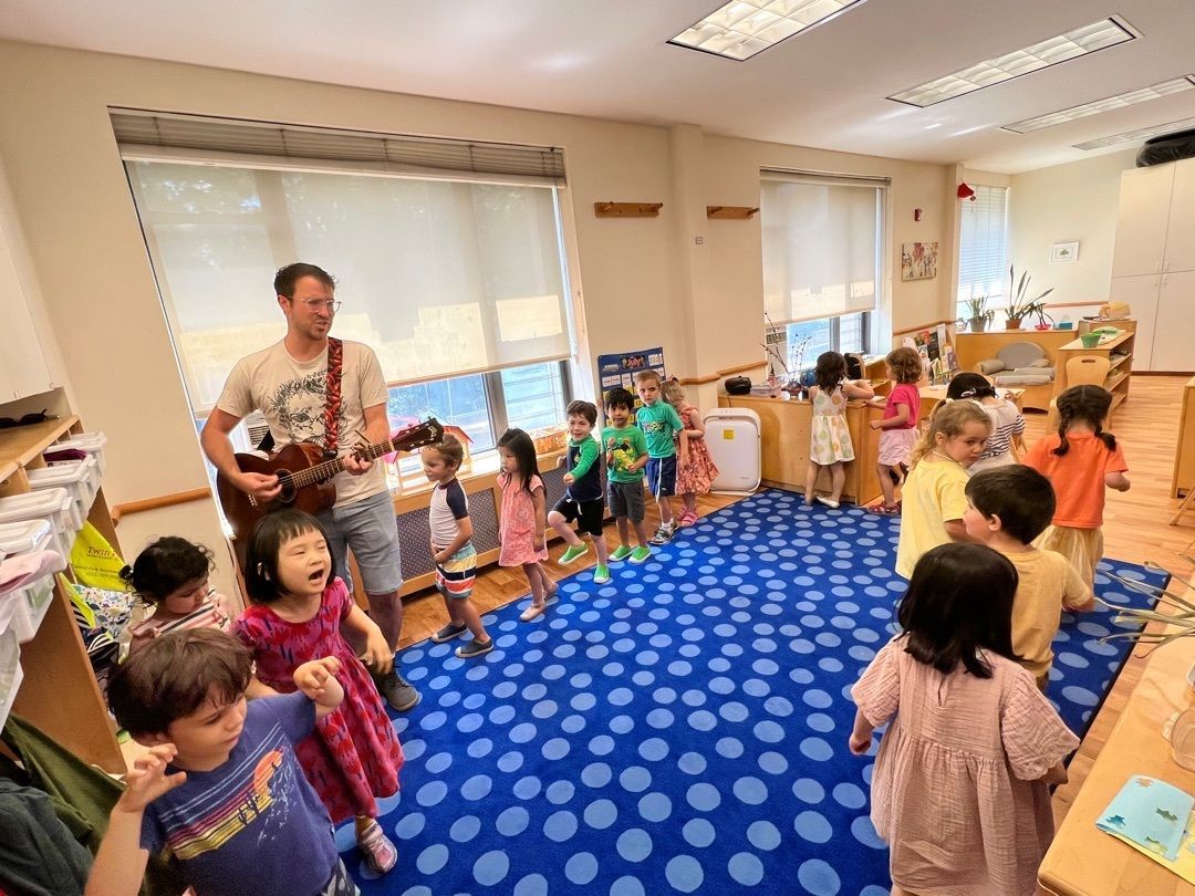 The Montessori music teacher with guitar sings to children in a classroom. Kids are singing and dancing on a blue carpet.
