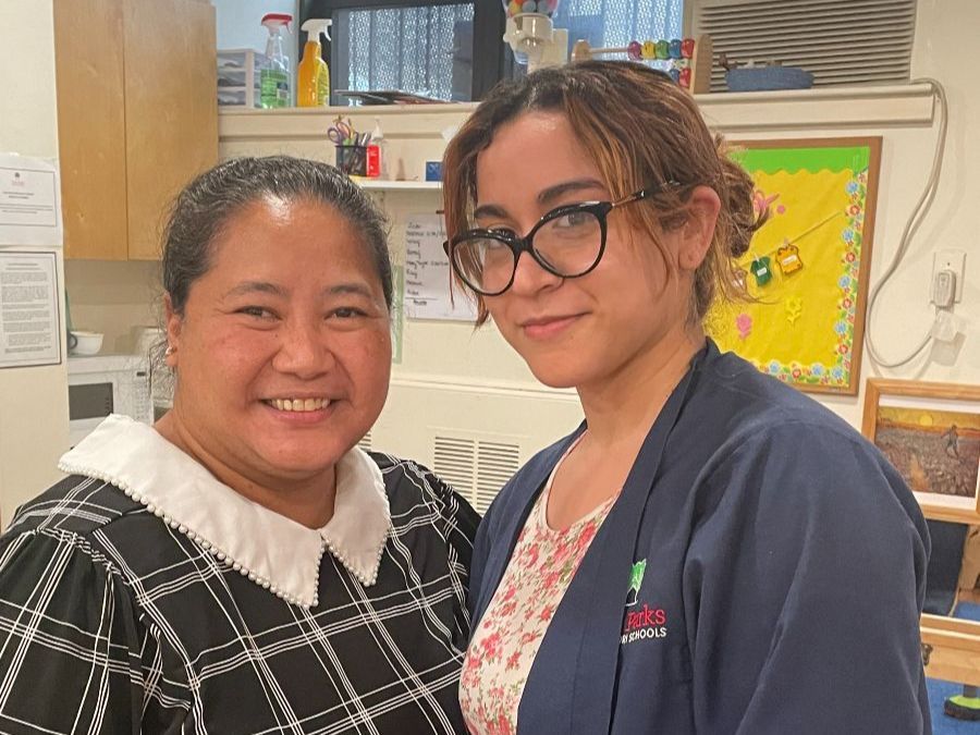 Two women smiling indoors: one in a patterned dress, the other in glasses and a navy jacket.