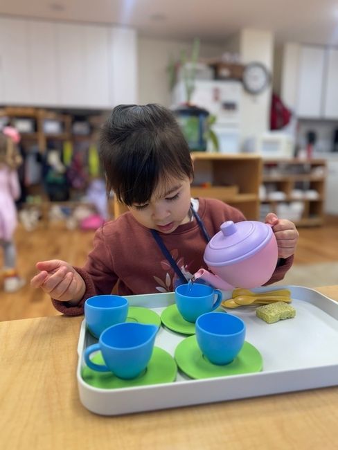 Child pouring from a purple pitcher into blue cups at a play table with toy dishes