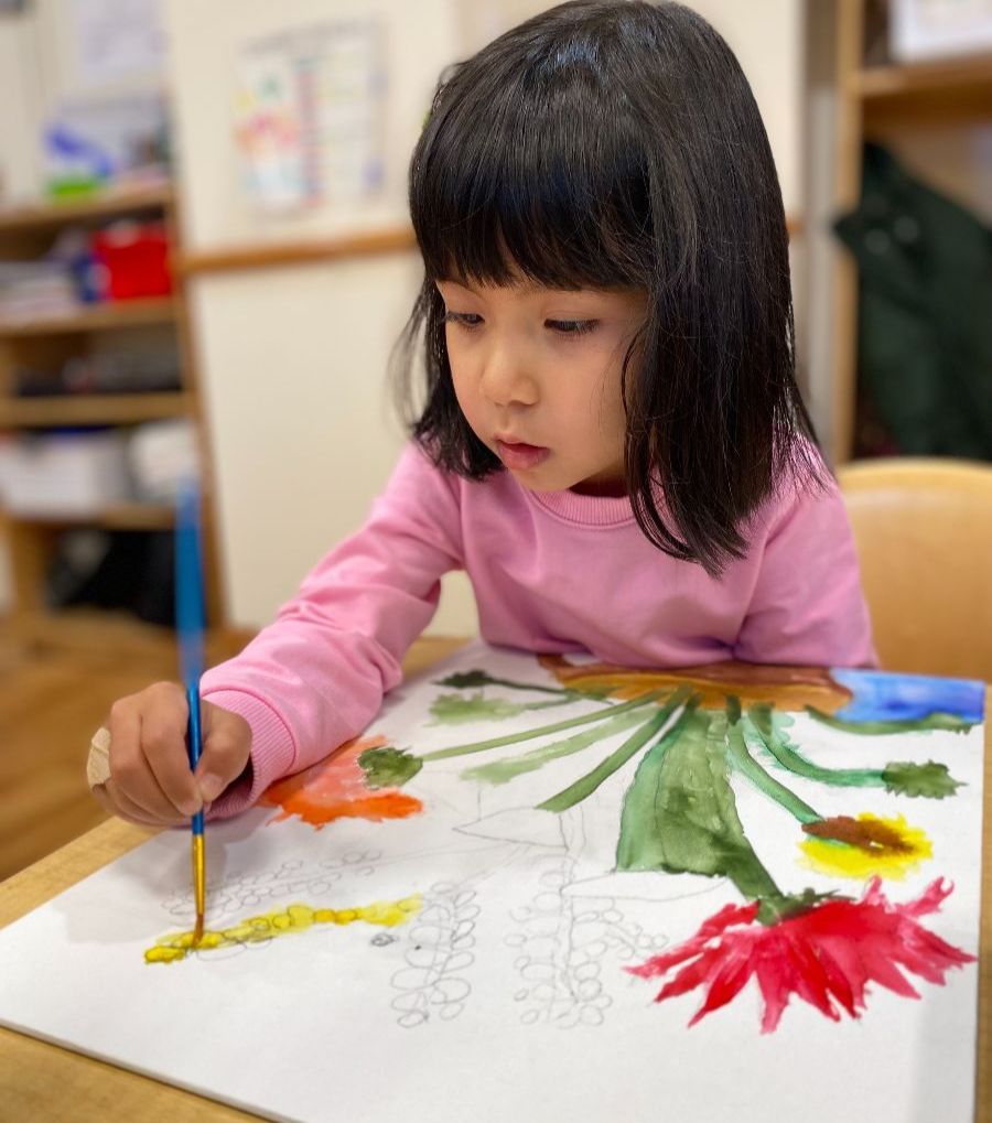 Montessori Child painting with watercolor, wearing pink, focused on her art, in a brightly lit room.