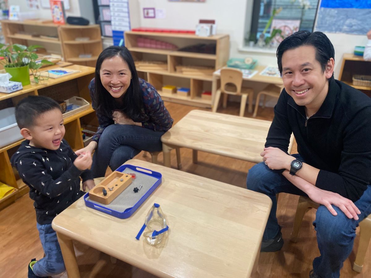A young child, a woman, and a man smile, watching a marble roll on a table in a classroom.