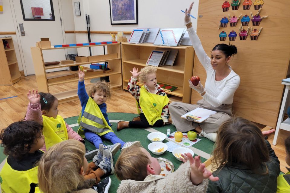 Children and teacher in a classroom circle; teacher holds an apple as kids raise their hands.