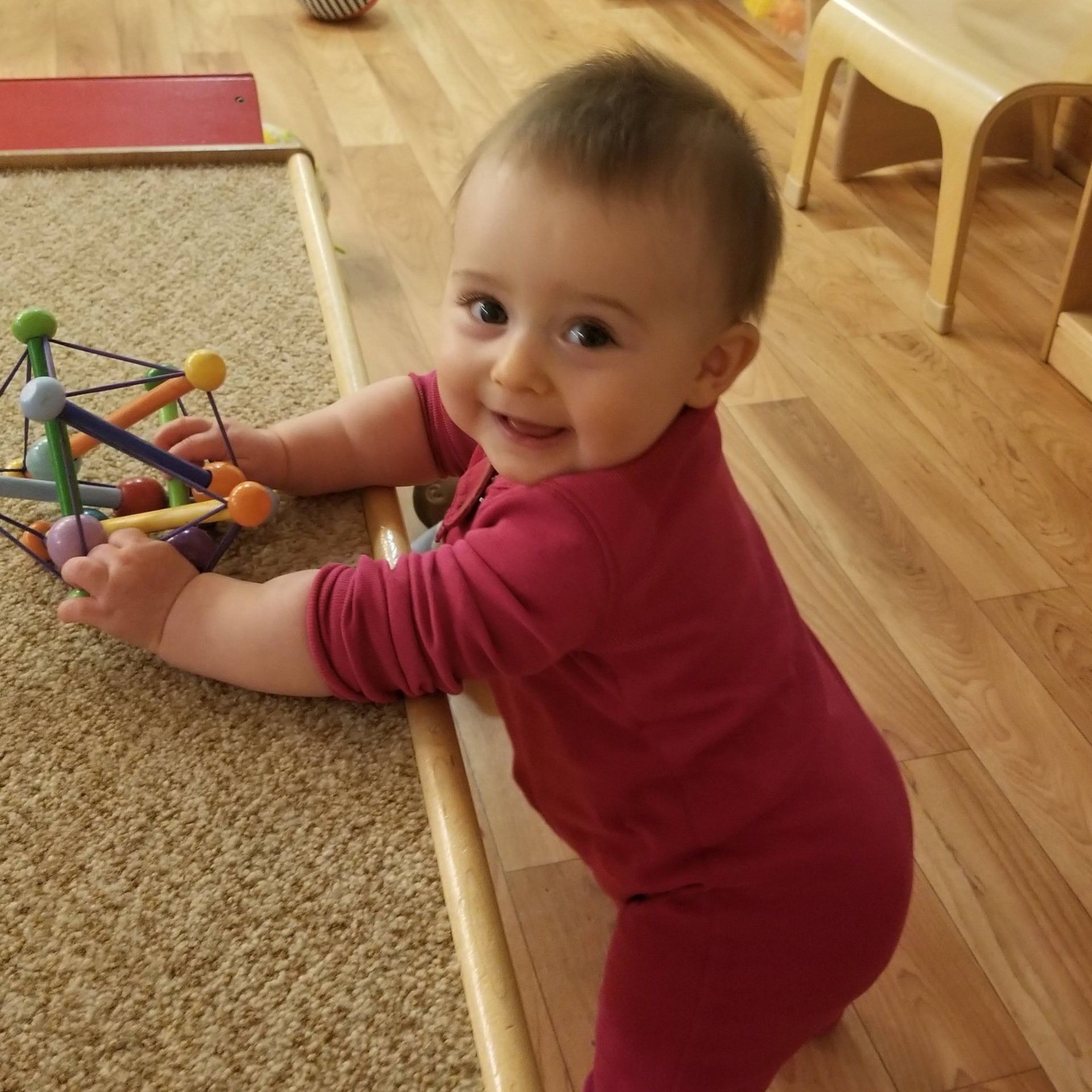 Smiling baby in red onesie playing on a wood floor, holding a colorful toy.
