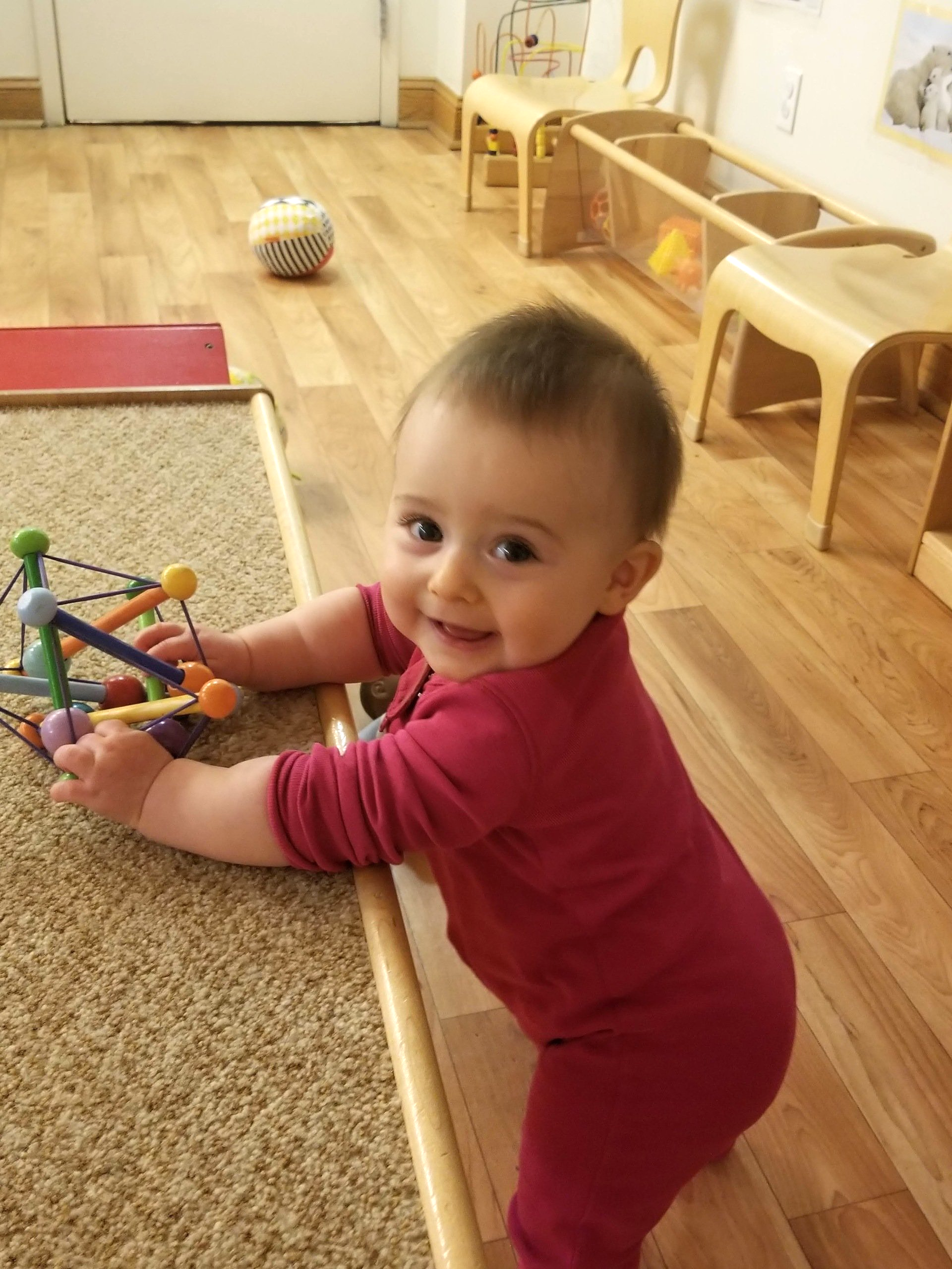 Smiling baby in red jumpsuit, holding a toy, standing on a wooden floor.