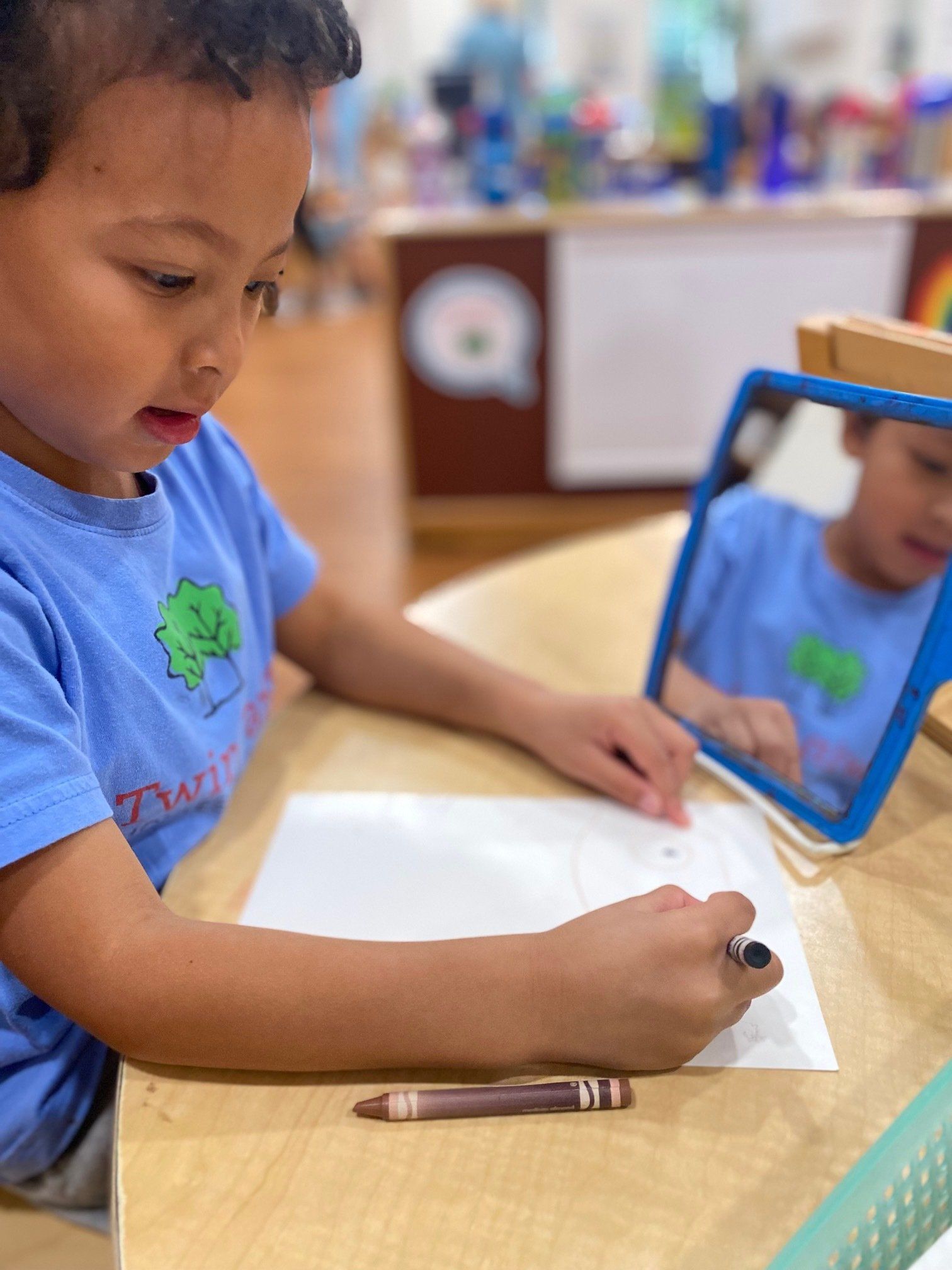 Boy in blue shirt draws with crayon, using mirror, at a table.
