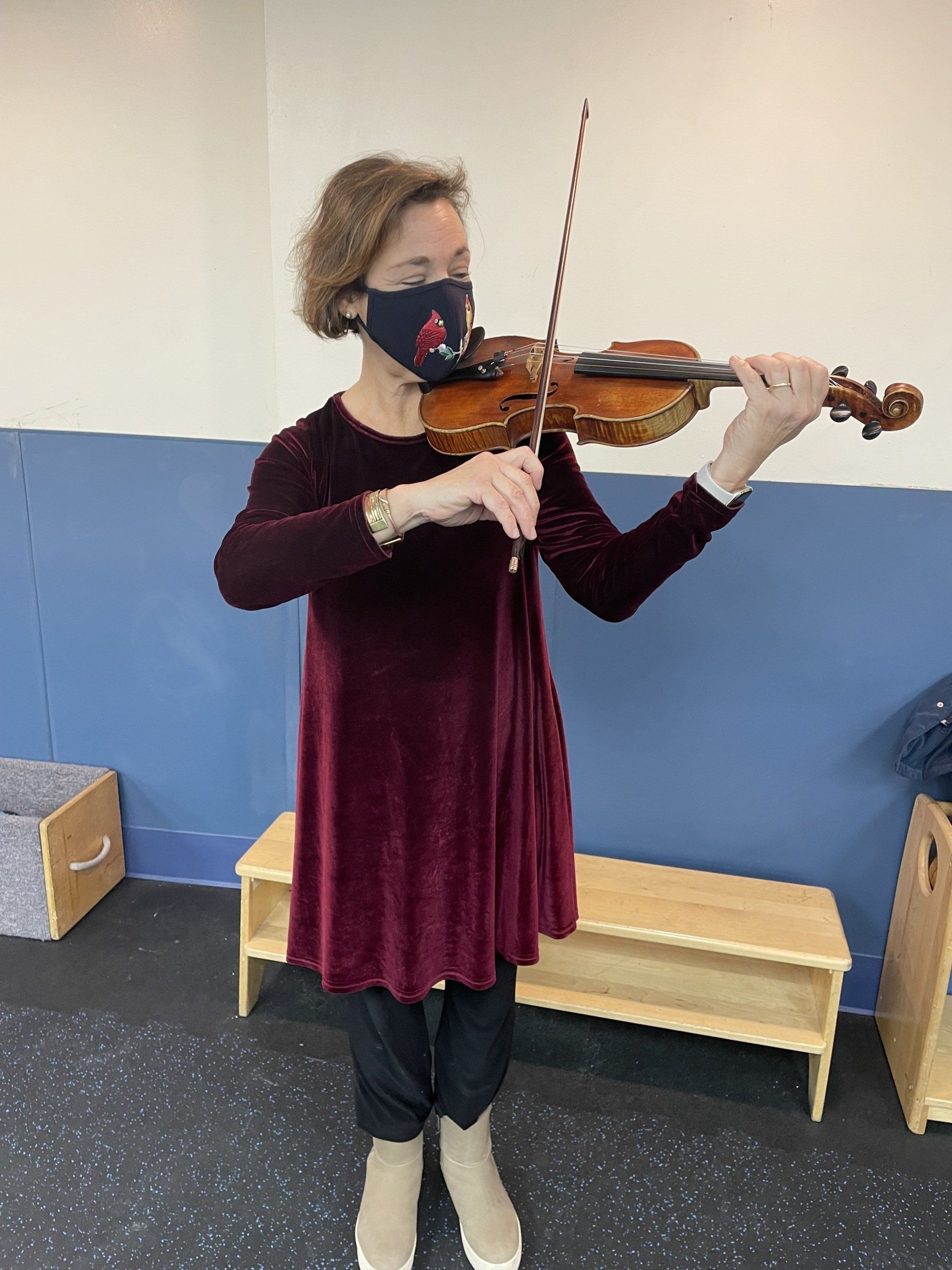 Woman playing a violin indoors, wearing a face mask and maroon dress; blue wall in the background.