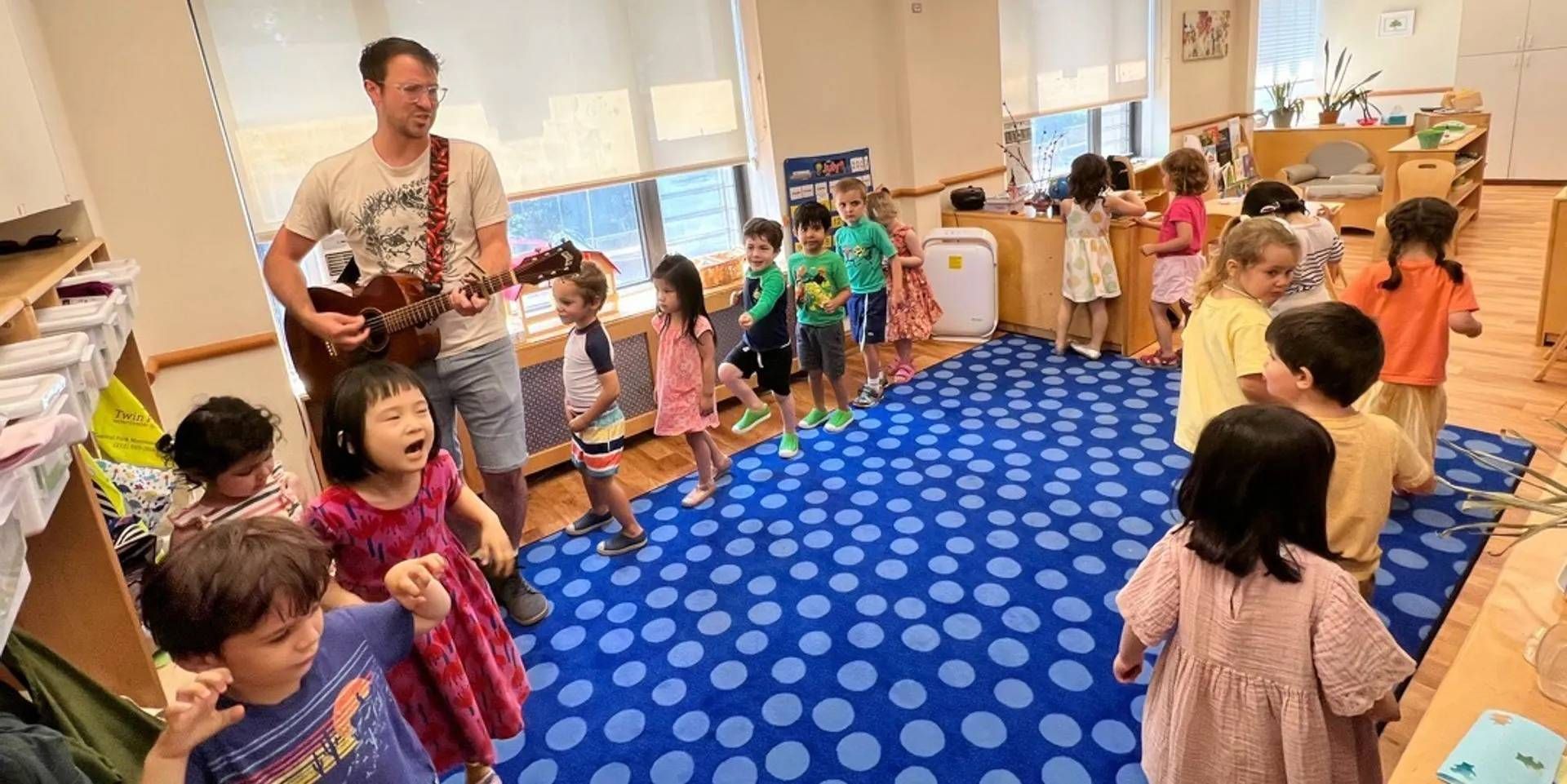 A music teacher plays guitar in a classroom surrounded by children singing and dancing on a blue polka dot rug.